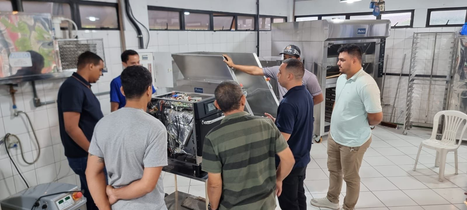 Um grupo de homens inspeciona equipamentos em uma cozinha industrial. Um dos homens aponta para a máquina.