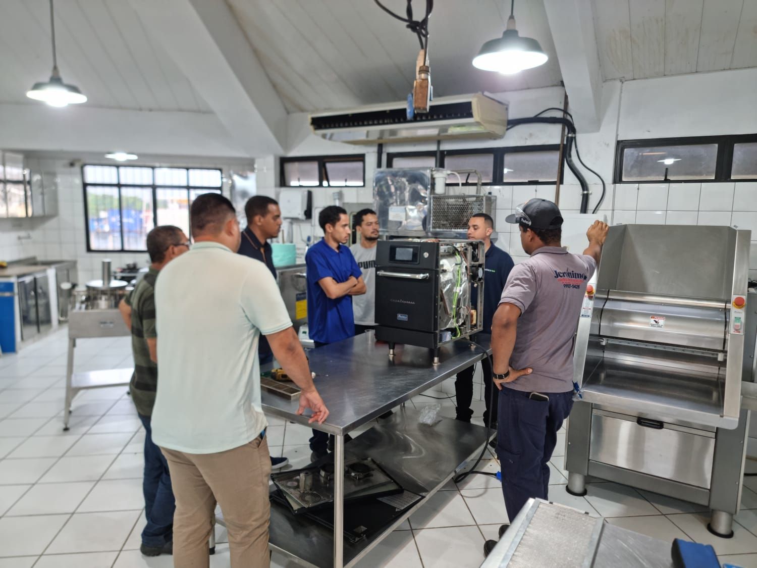 Grupo de homens observando um equipamento sobre uma mesa de aço inoxidável em uma cozinha.