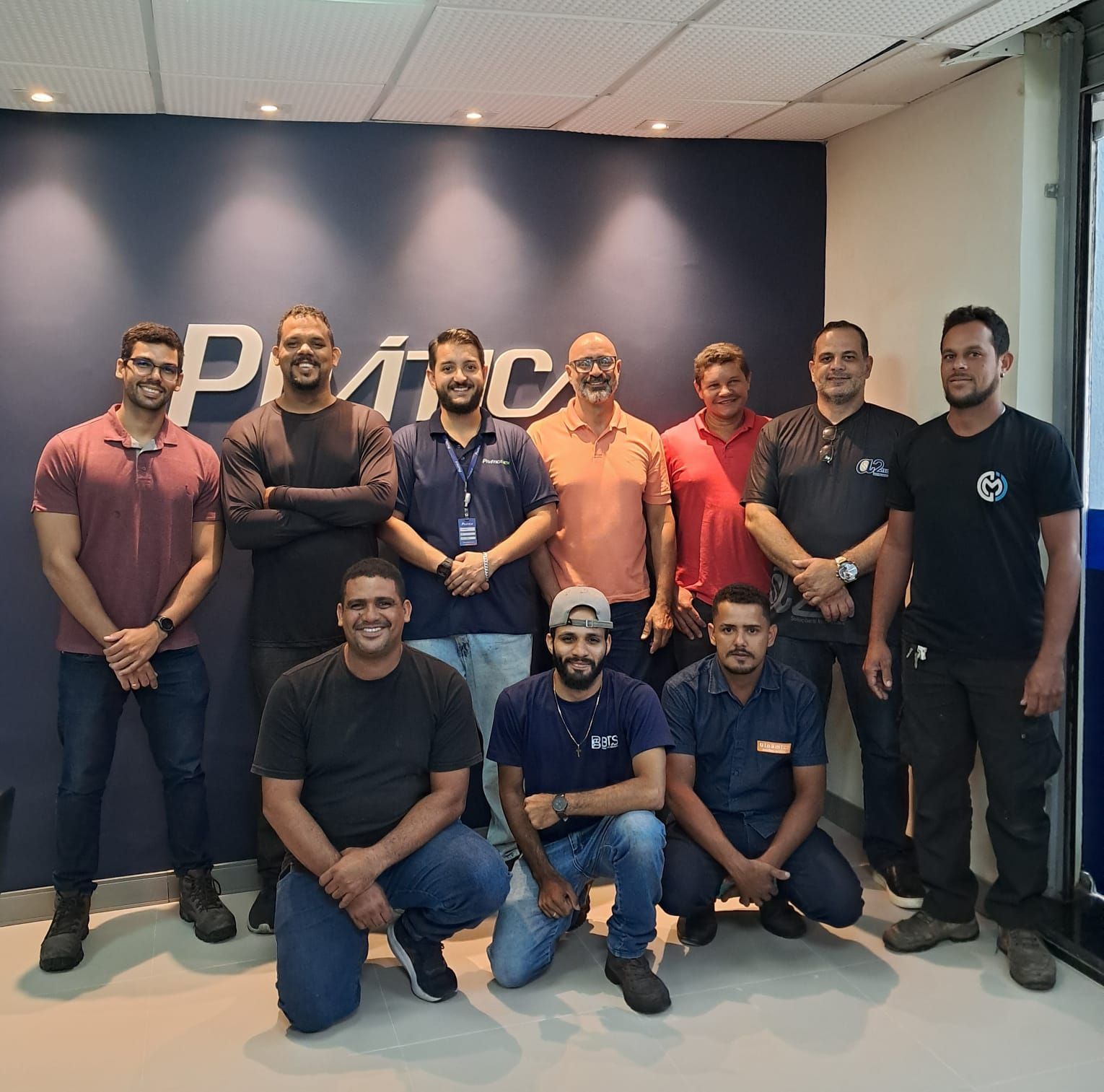 Grupo de pessoas posando em frente a uma parede com o logotipo Prática