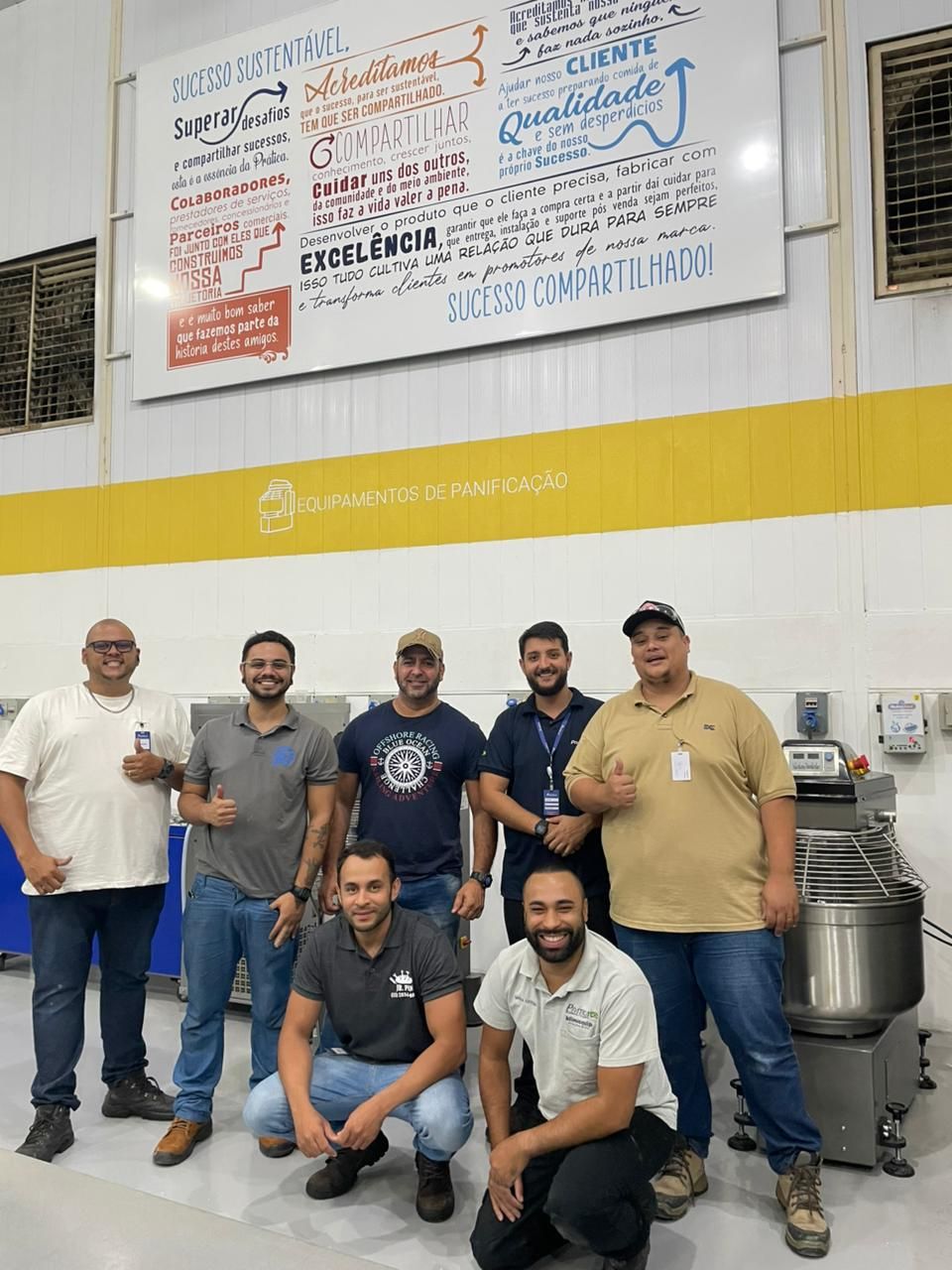Grupo de homens em um ambiente fabril, posando perto de equipamentos e um quadro branco com texto.