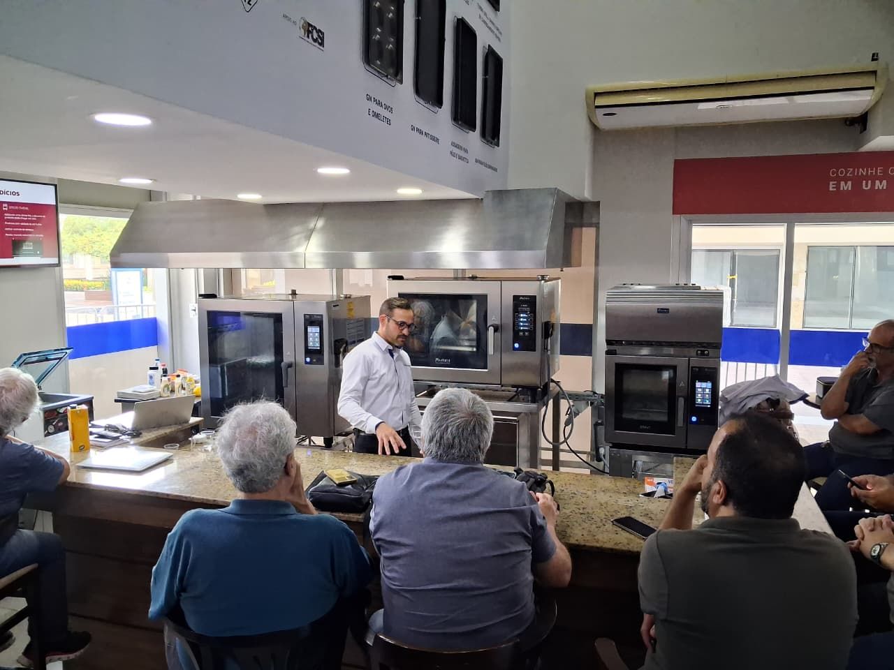 Um chef demonstrando o funcionamento de fornos para um grupo de homens em uma cozinha.