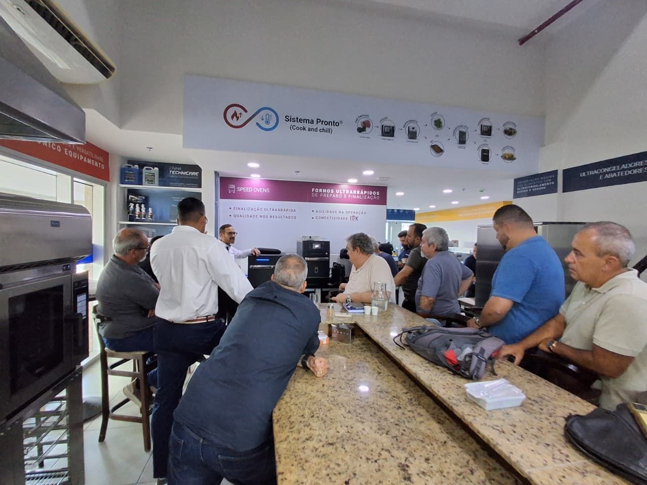 Grupo de homens em um balcão, possivelmente de uma loja ou local de reunião, com produtos expostos e uma placa.