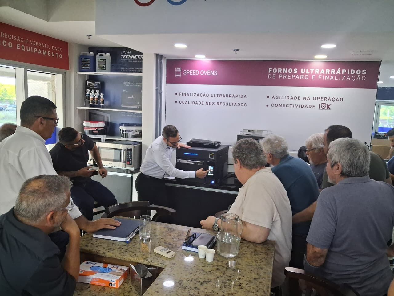 Pessoas se reuniram para assistir a um homem demonstrando aparelhos eletrônicos em uma loja com sinalização.