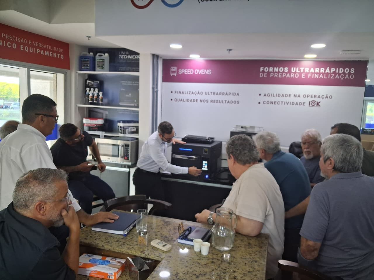 Um homem demonstra um aparelho de som para um grupo de homens em uma loja, com eletrodomésticos ao fundo.