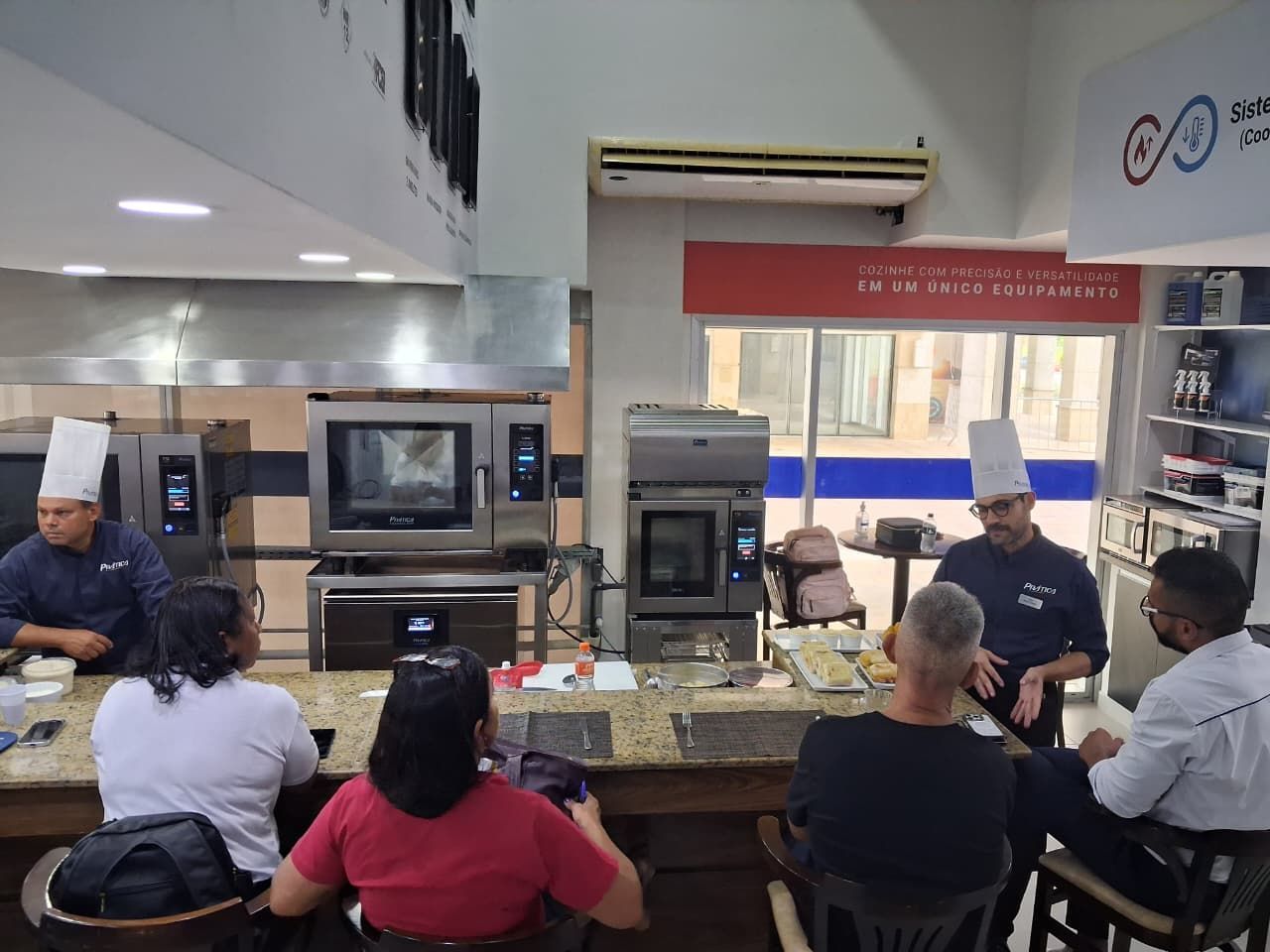 Chefs em uma cozinha demonstrando técnicas culinárias. Os participantes observam em um ambiente de aula de culinária.
