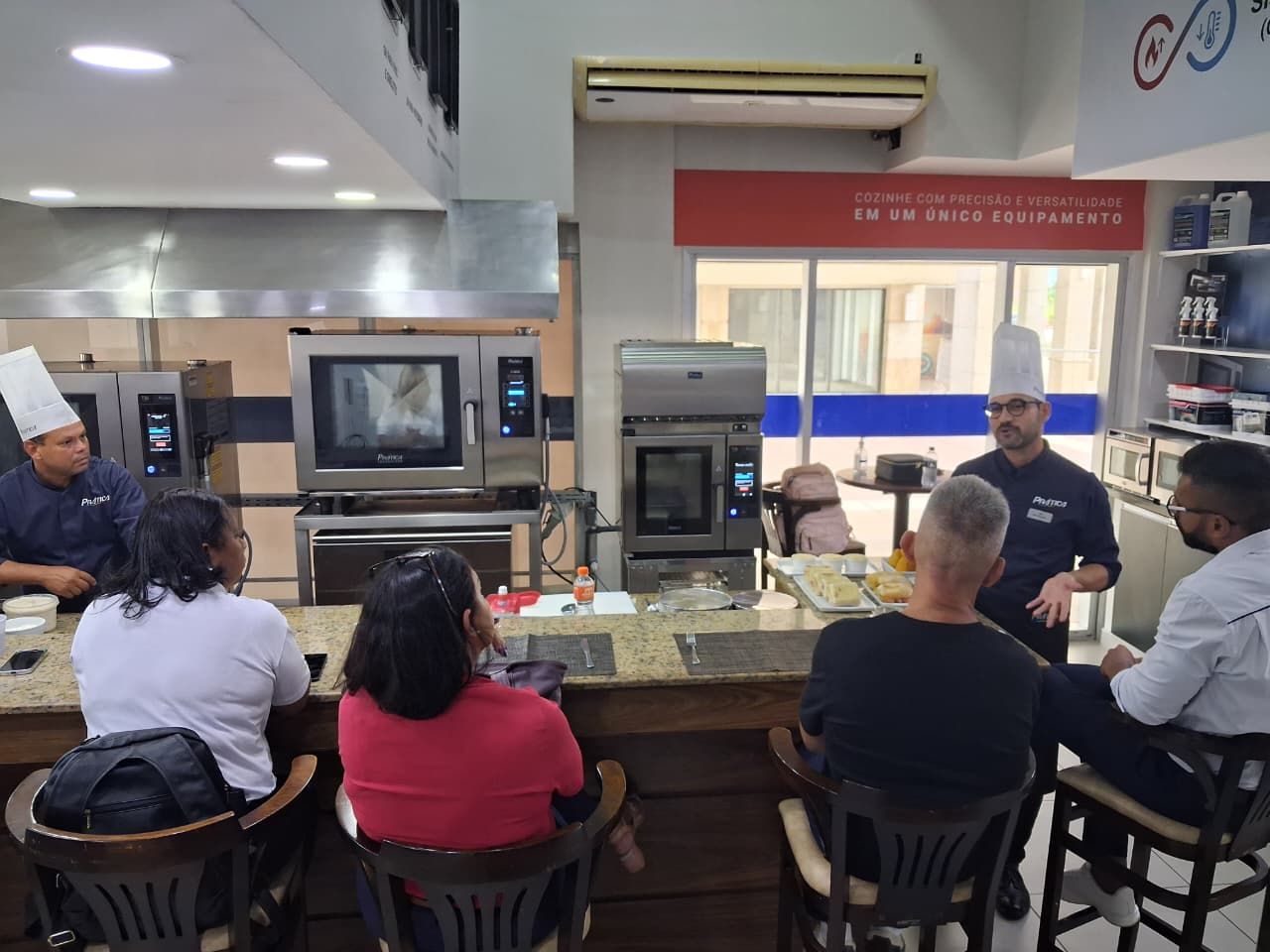 Um chef instruindo um pequeno grupo em uma cozinha com fornos. O grupo está sentado em um balcão, observando.