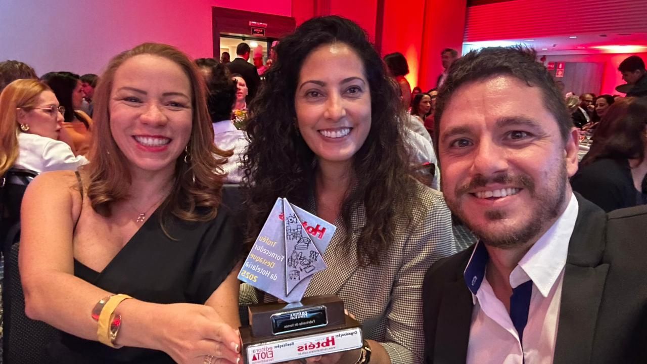 Três pessoas sorrindo, segurando um prêmio em uma sala de eventos bem iluminada.