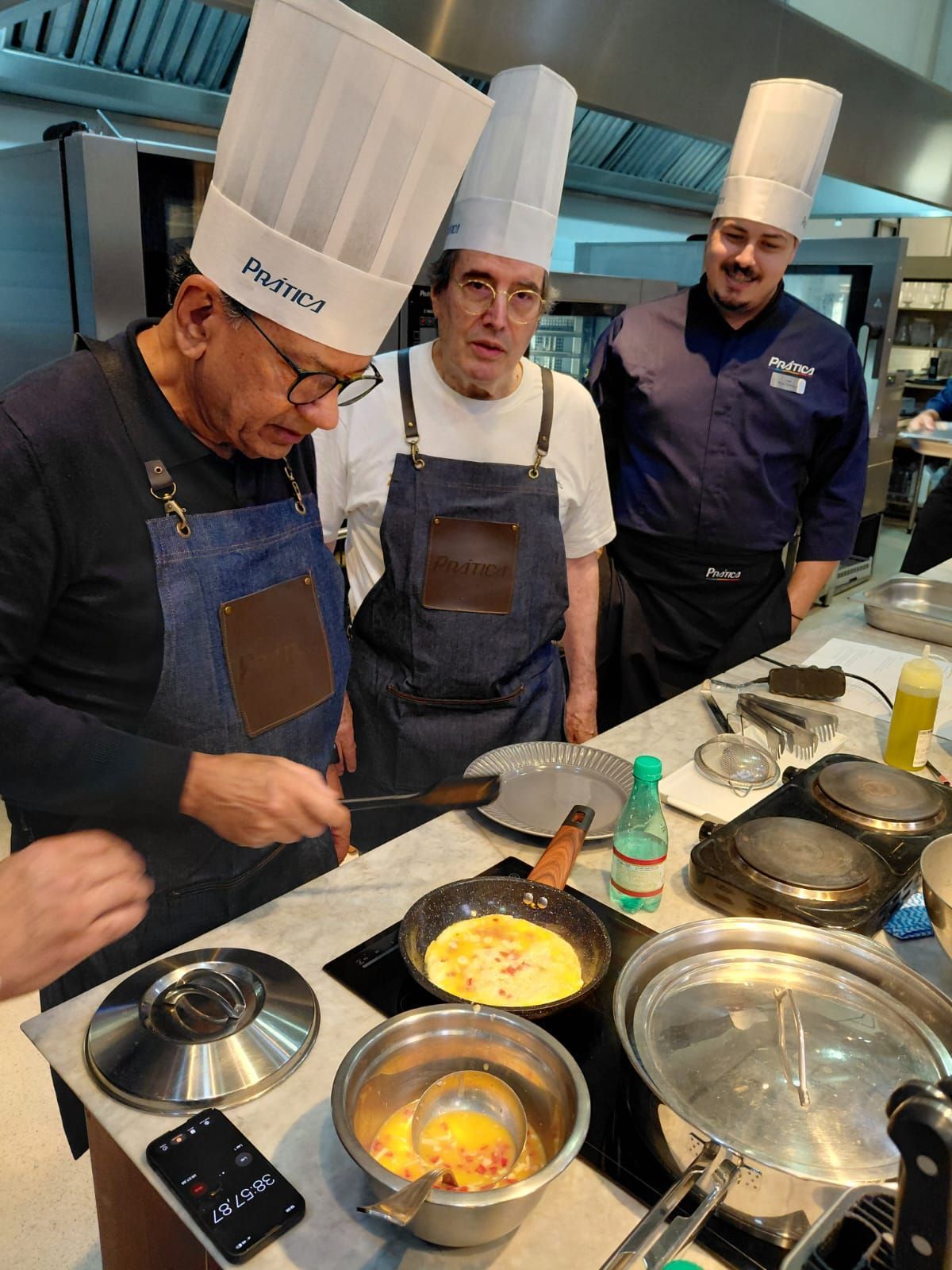 Chefs de chapéu cozinhando em uma cozinha. Um deles vira a comida em uma panela enquanto os outros observam.