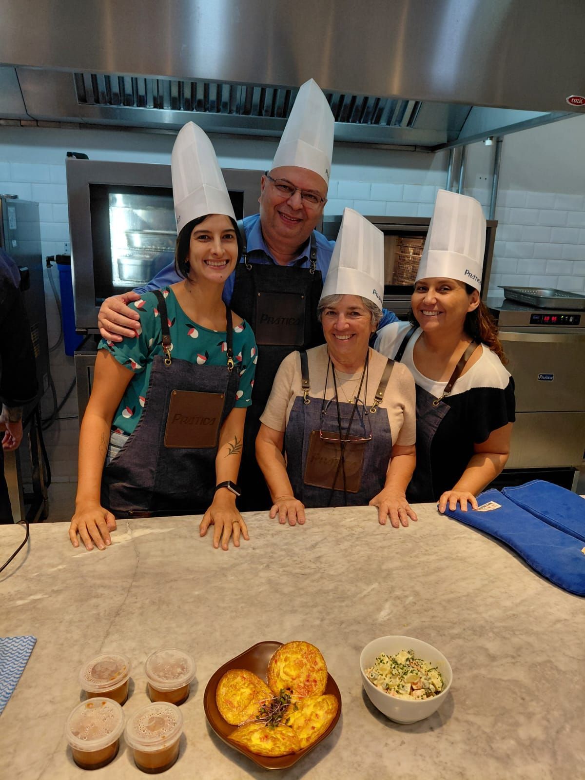Quatro pessoas com chapéus e aventais de chef posam em uma cozinha, sorrindo enquanto observam a comida preparada.
