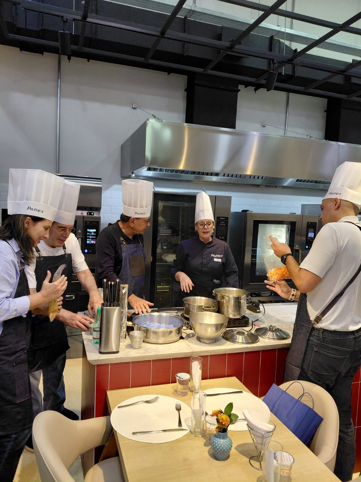 Chefs com chapéus altos cozinham em uma cozinha profissional, observando uma demonstração, com os equipamentos ao fundo.