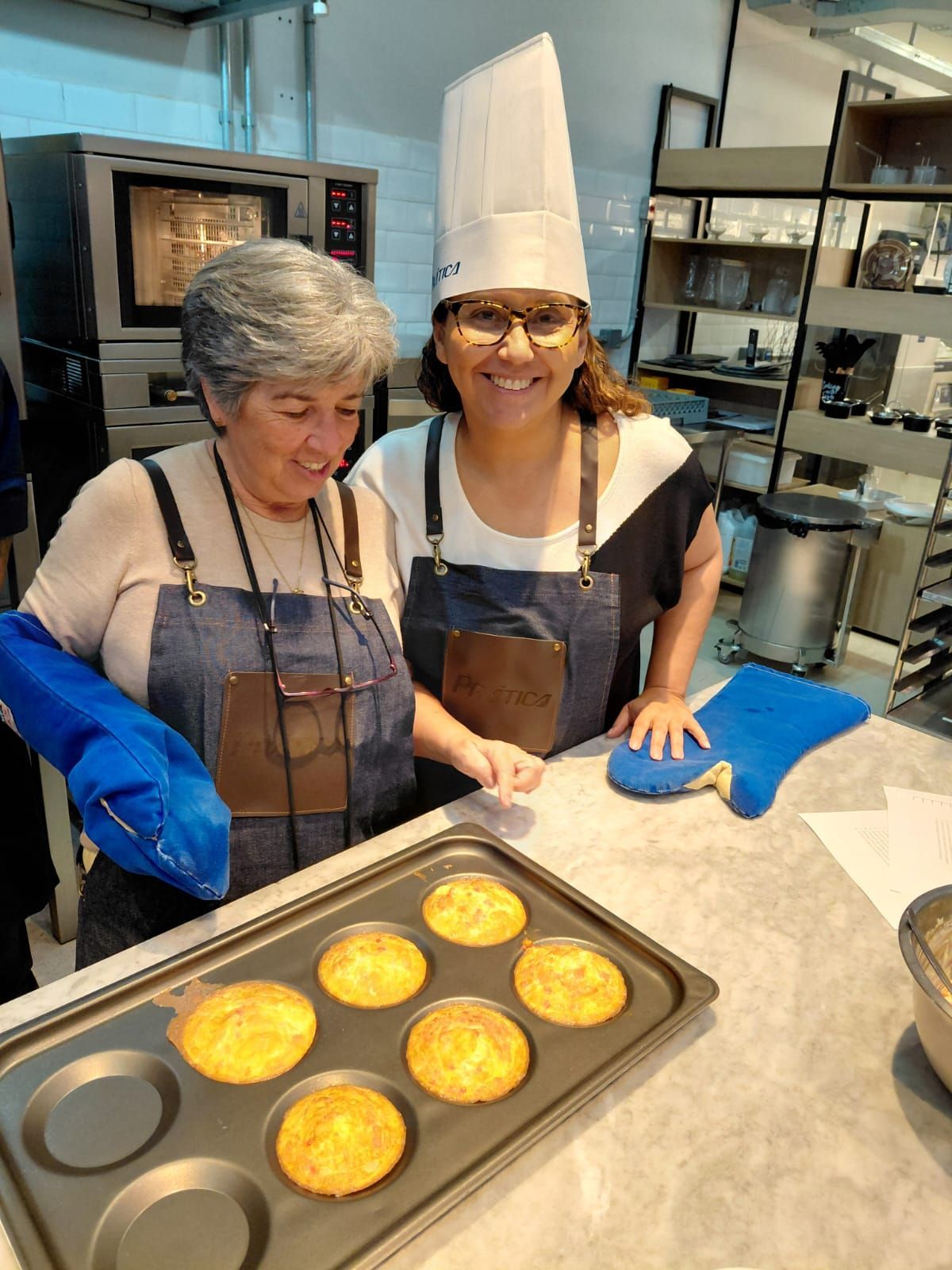 Duas pessoas de avental e luvas de forno com uma bandeja de muffins, em um ambiente de cozinha.
