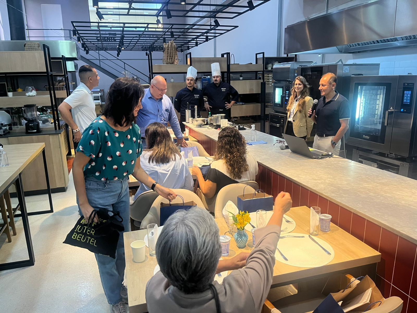 Uma demonstração culinária: chefs e participantes em uma cozinha moderna com uma grande bancada, fornos e mesas.