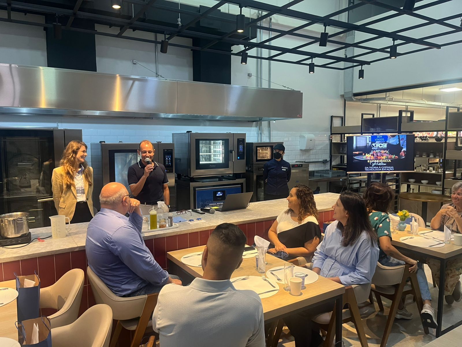 Pessoas sentadas à mesa assistem a uma apresentação em uma cozinha. O apresentador segura um microfone.