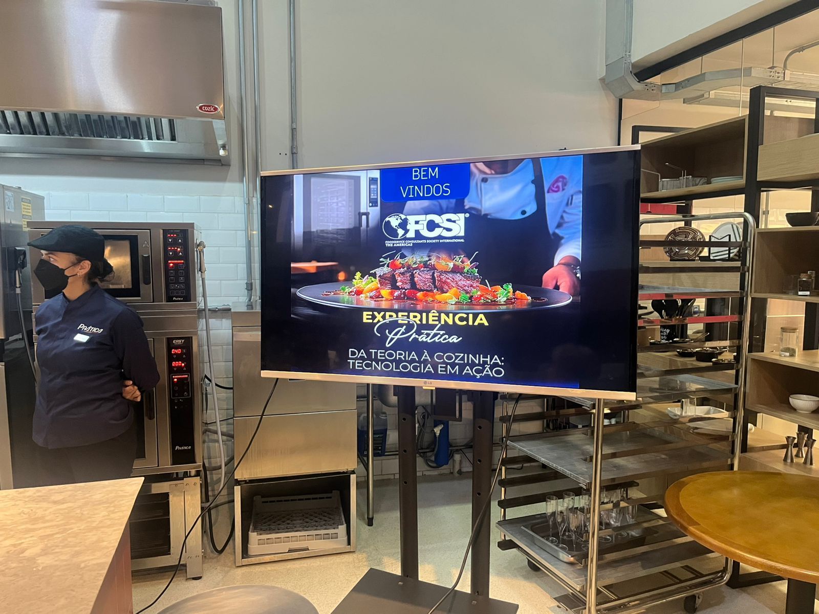 Mulher usando máscara facial está perto de fornos e de uma TV que exibe um programa de culinária. Ambiente de cozinha.