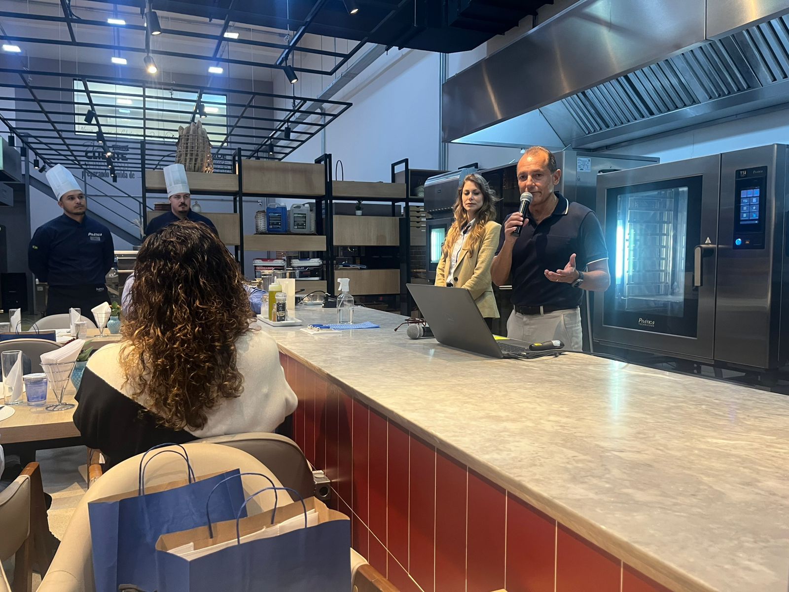 Homem falando ao microfone, em pé atrás de um balcão com uma mulher, chefs ao fundo.