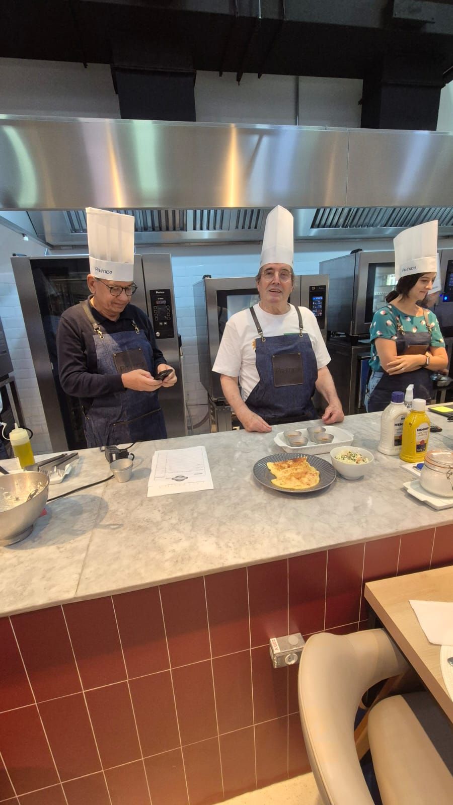 Chefs de chapéu e avental brancos posam atrás de um balcão na cozinha, sorrindo para a câmera.