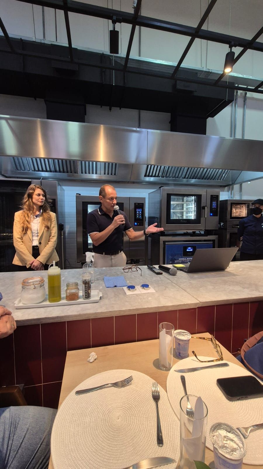 Homem falando em um balcão, mulher ao lado dele, cozinha profissional ao fundo.