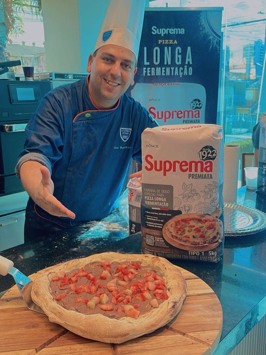 Um homem está de pé ao lado de uma pizza em uma tábua de corte de madeira.