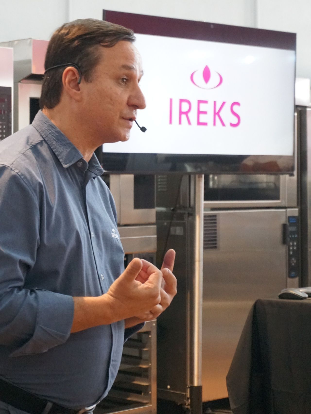 Homem falando em frente a uma tela com o logotipo da IREKS. Ele veste uma camisa azul e gesticula com as mãos em uma cozinha.
