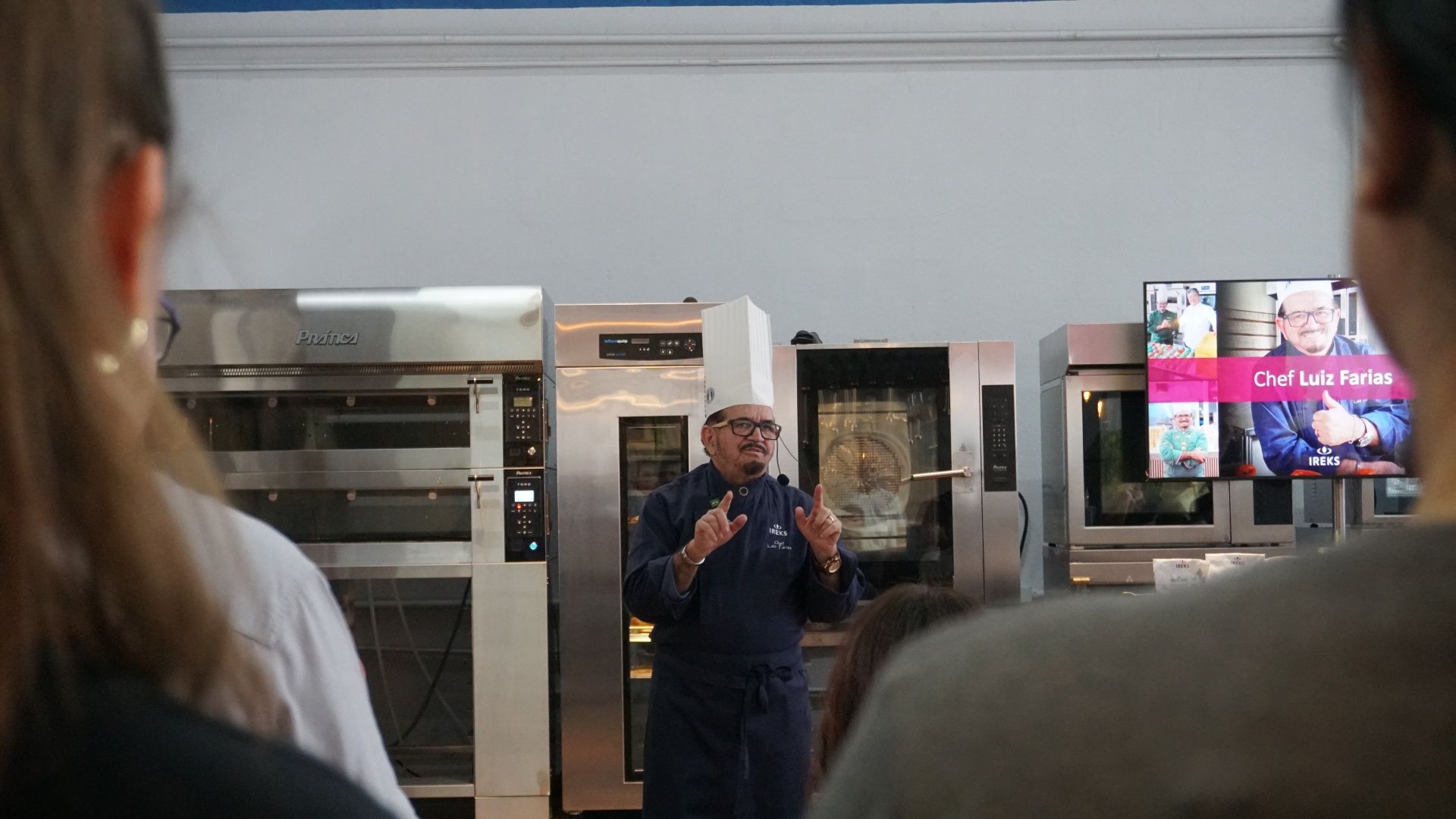 Chef falando em evento em frente a fornos; público em primeiro plano.