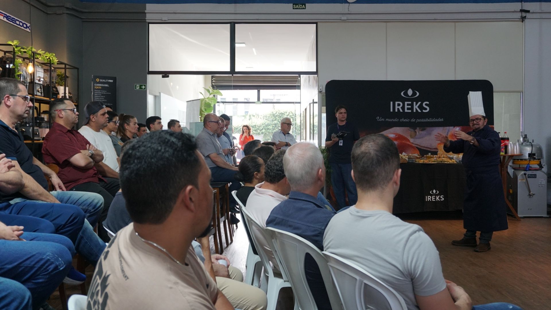 Chef fazendo uma demonstração para uma plateia sentada; cenário com a marca IREKS.