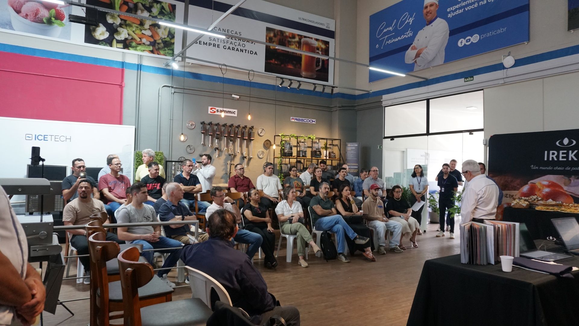 Um chef fazendo uma apresentação para uma plateia em um workshop. Diversos utensílios de cozinha e alimentos expostos.