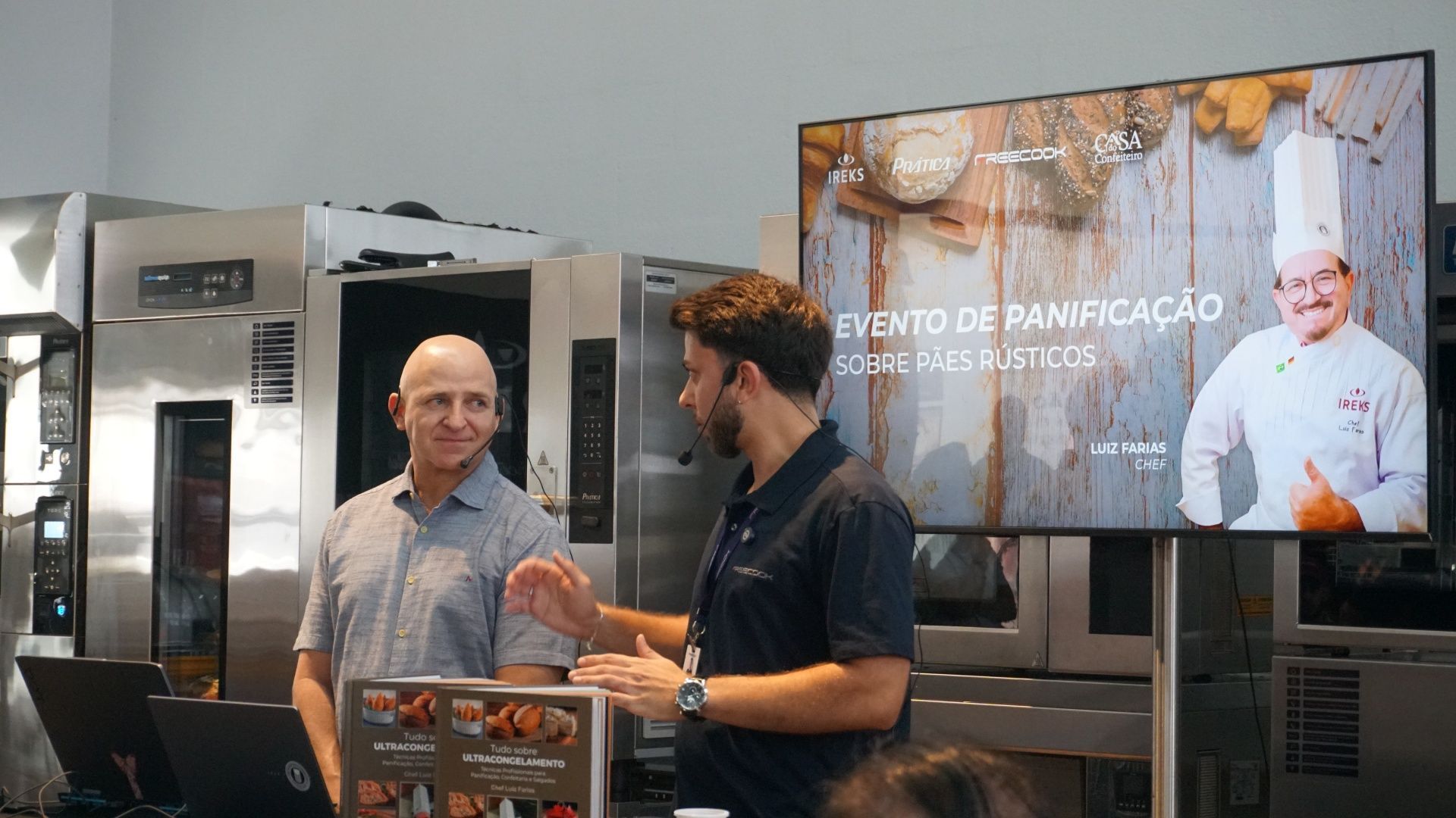 Dois homens em um evento de confeitaria, um deles gesticulando perto de uma tela com a imagem de um chef. Fornos ao fundo.