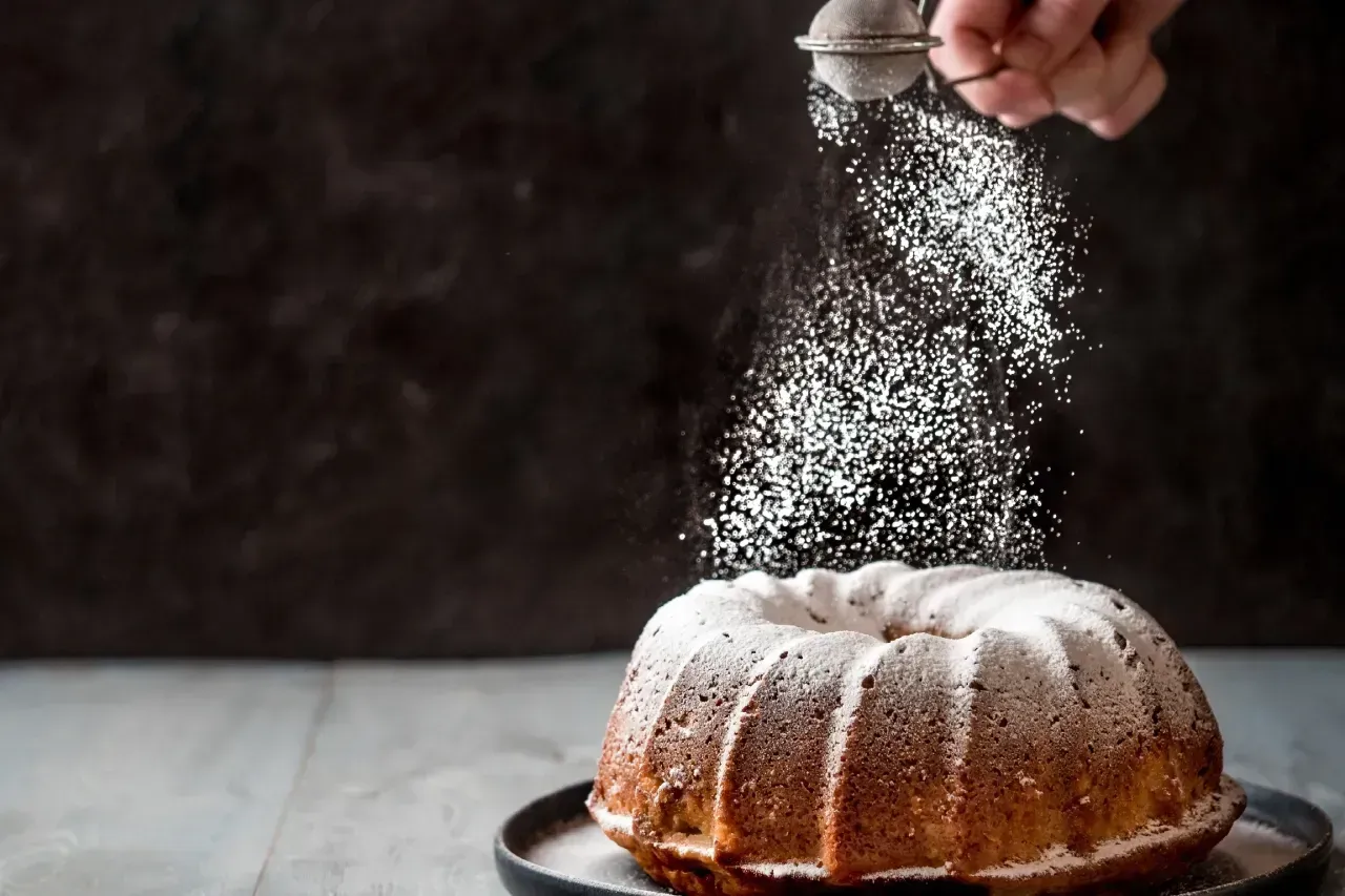 Uma mão polvilha açúcar de confeiteiro sobre um bolo Bundt contra um fundo escuro e rústico.