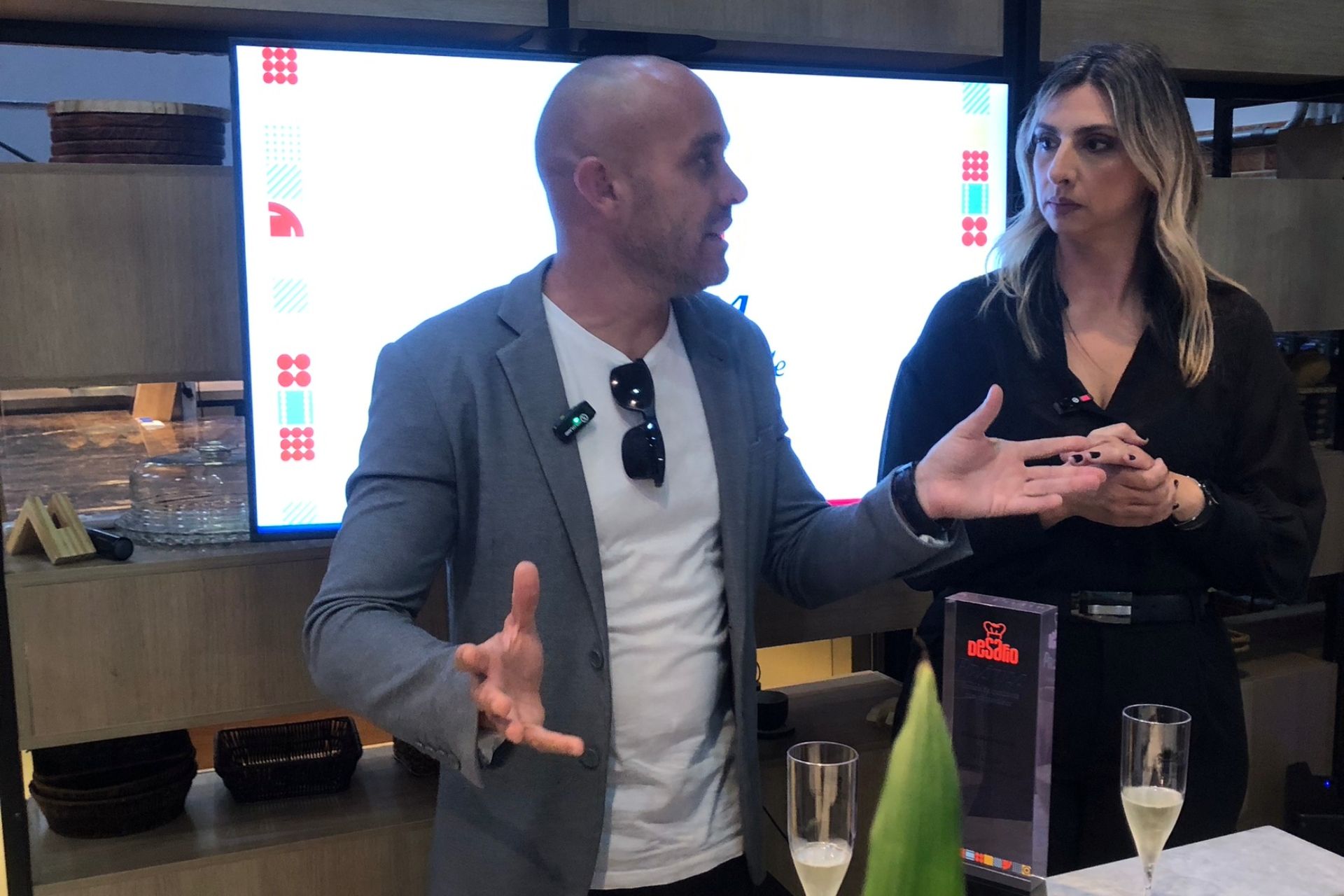 Homem gesticula, fala; mulher ouve. Ambos estão em frente a uma tela. Prêmios e taças de champanhe em primeiro plano.