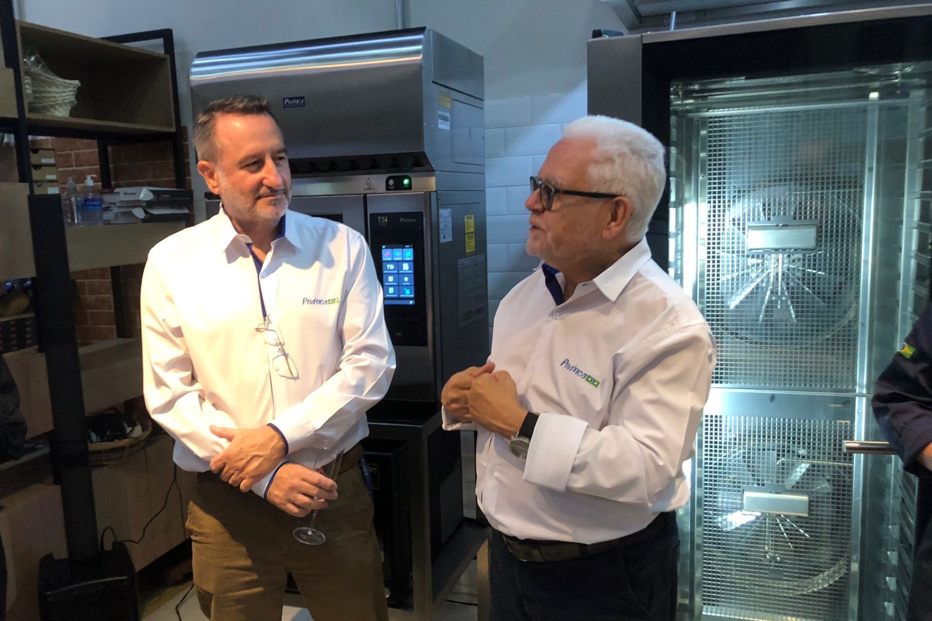 Dois homens de camisa branca conversando ao lado de fornos industriais em uma cozinha comercial.