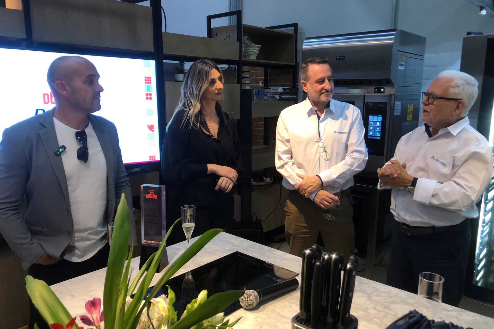 Quatro pessoas estão em pé junto a uma bancada em uma cozinha moderna. Um homem fala, gesticulando; os outros escutam.