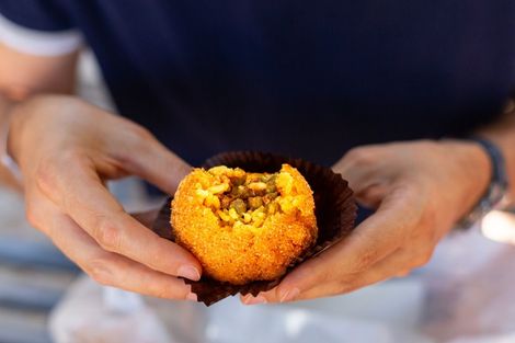 Mãos segurando um arancini amarelo mordido, envolto em uma embalagem marrom, sobre um fundo de mesa desfocado.