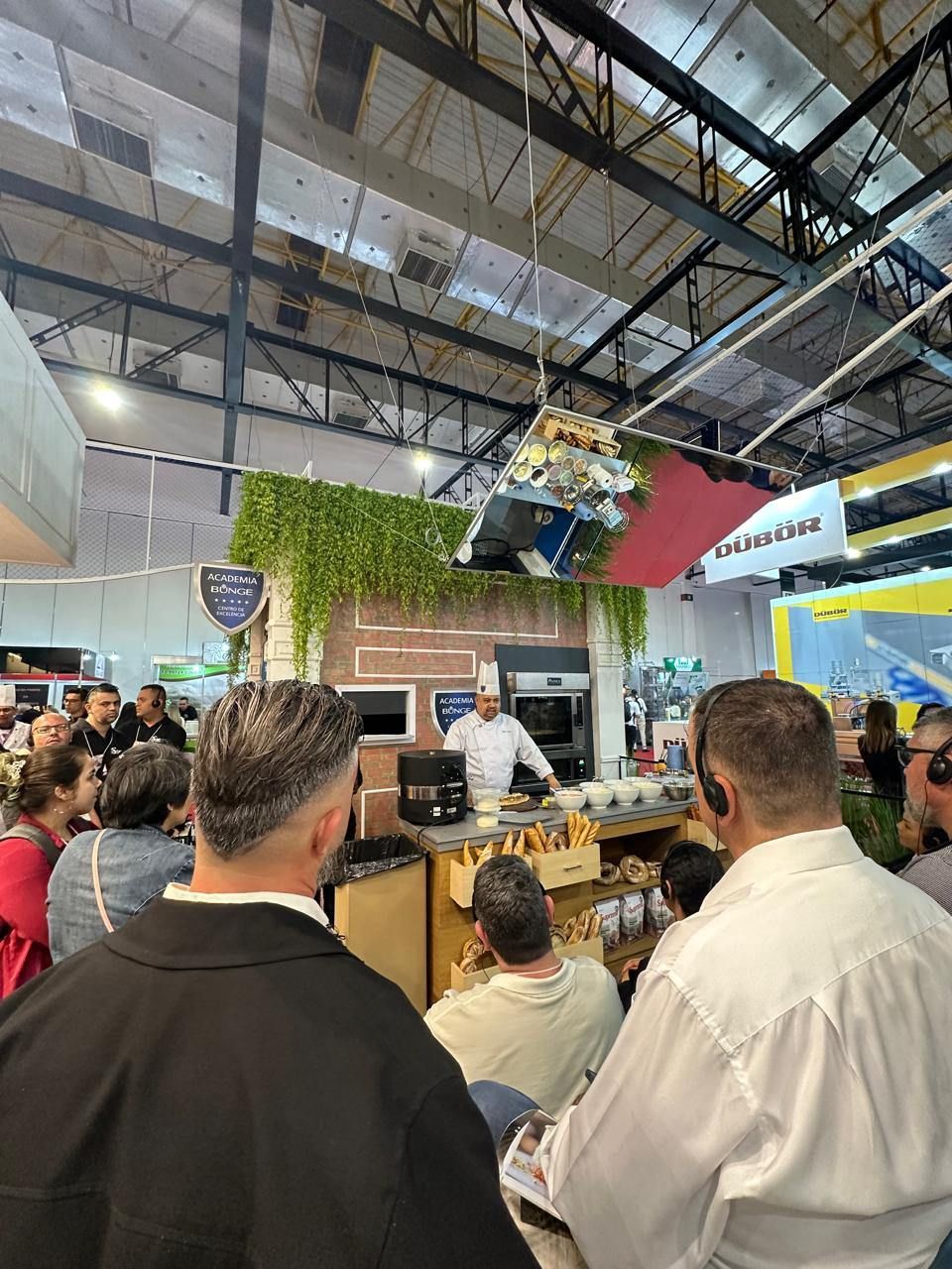 Um grupo de pessoas está sentado em uma sala observando um homem cozinhar.