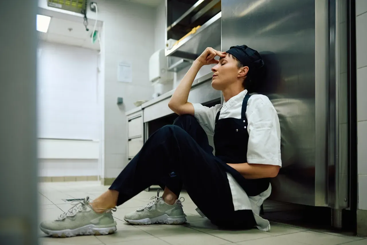 Uma pessoa vestida com uniforme de chef está sentada no chão, encostada em uma geladeira em uma cozinha comercial, com uma expressão de estresse.