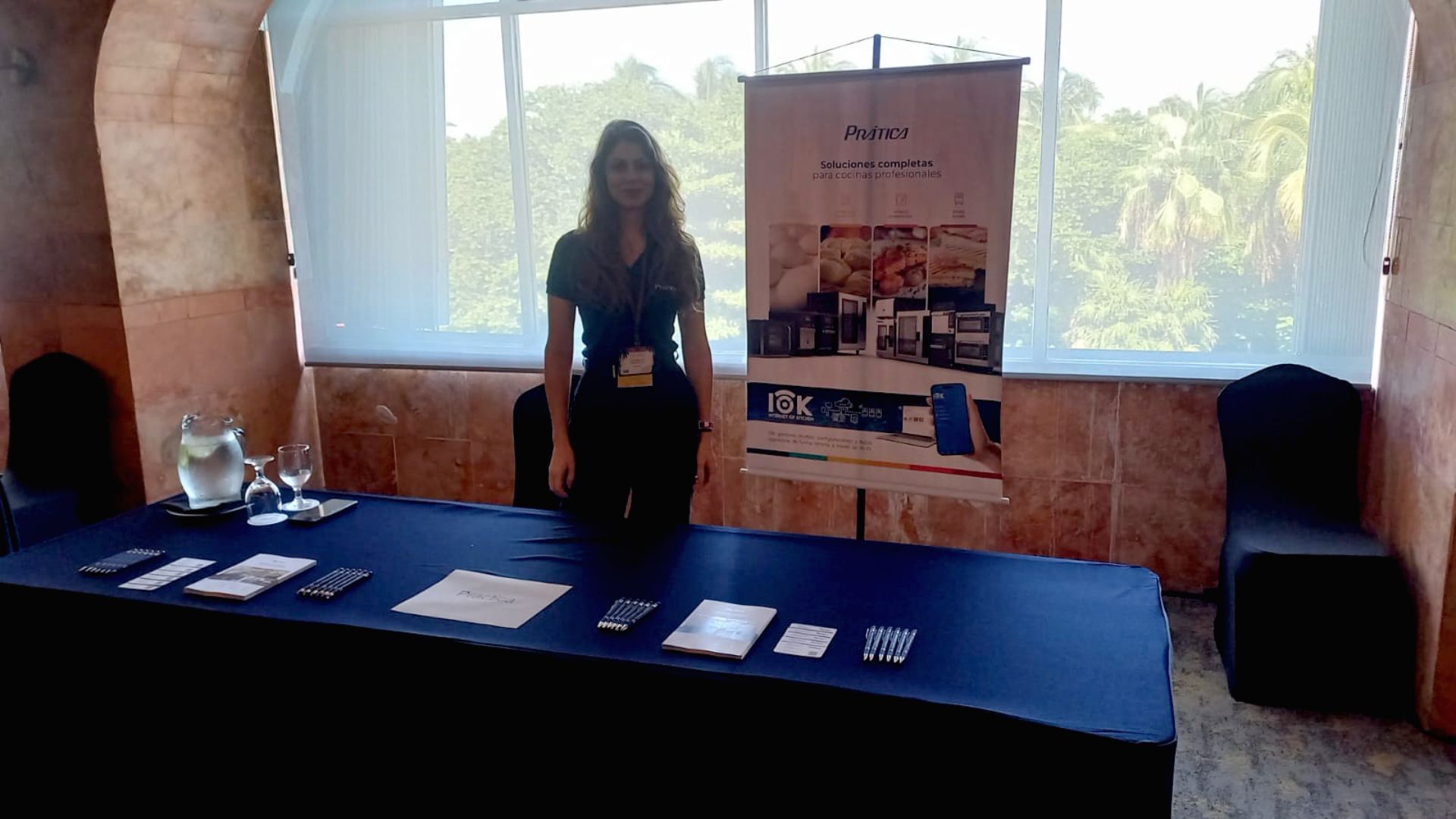 Mulher em pé atrás de uma vitrine, em frente a uma vitrine e um banner. Ela veste blusa e calça escuras.
