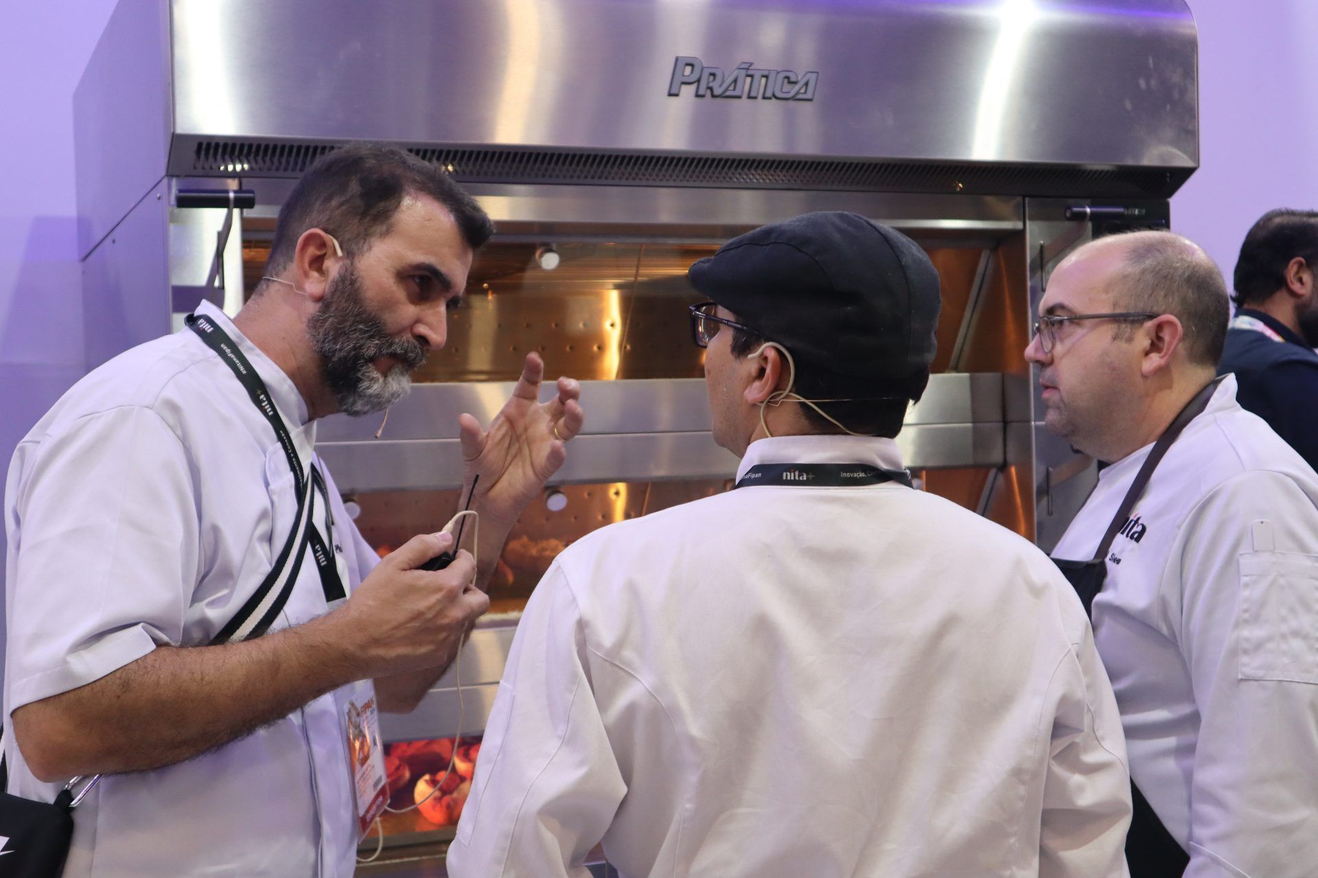 Um grupo de homens está em frente a um forno de aço inoxidável conversando entre si.