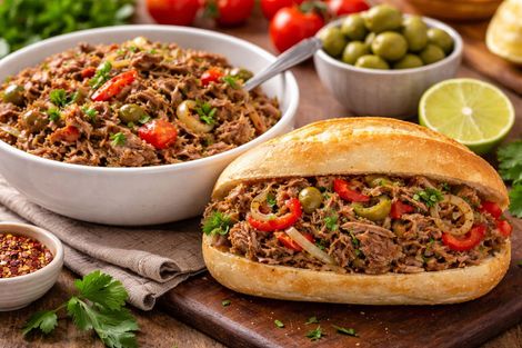 Sanduíche de carne louca em pão francês e vasilha de cerâmica com carne desfiada ao fundo.