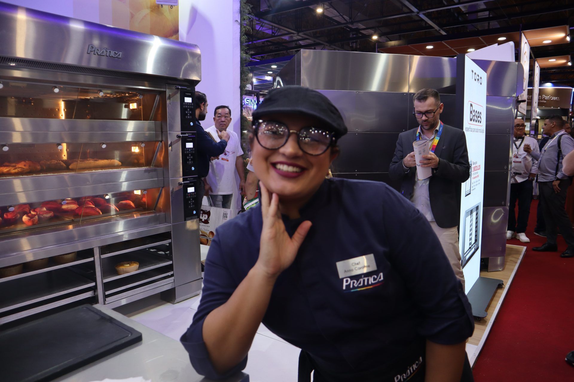 Uma mulher usando óculos e chapéu está sorrindo em frente a um forno.