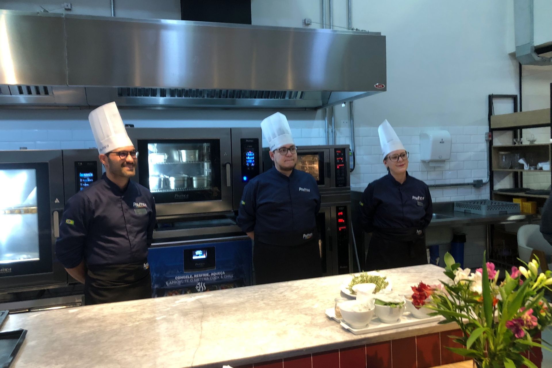 Três chefs com chapéus brancos e uniformes azuis estão em uma cozinha profissional.