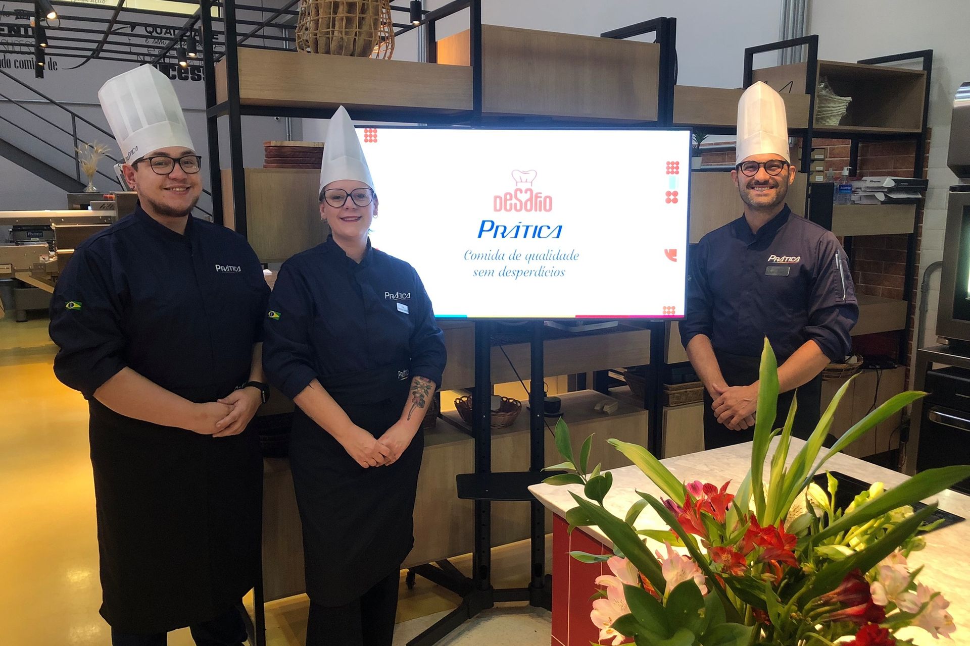 Três chefs de uniforme branco, em pé em frente a um monitor com um logotipo.