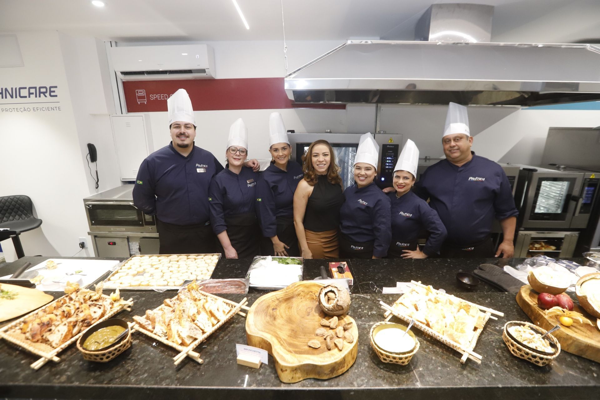 Chefs e uma mulher posam em uma cozinha comercial atrás de uma mesa de comida em um balcão preto.