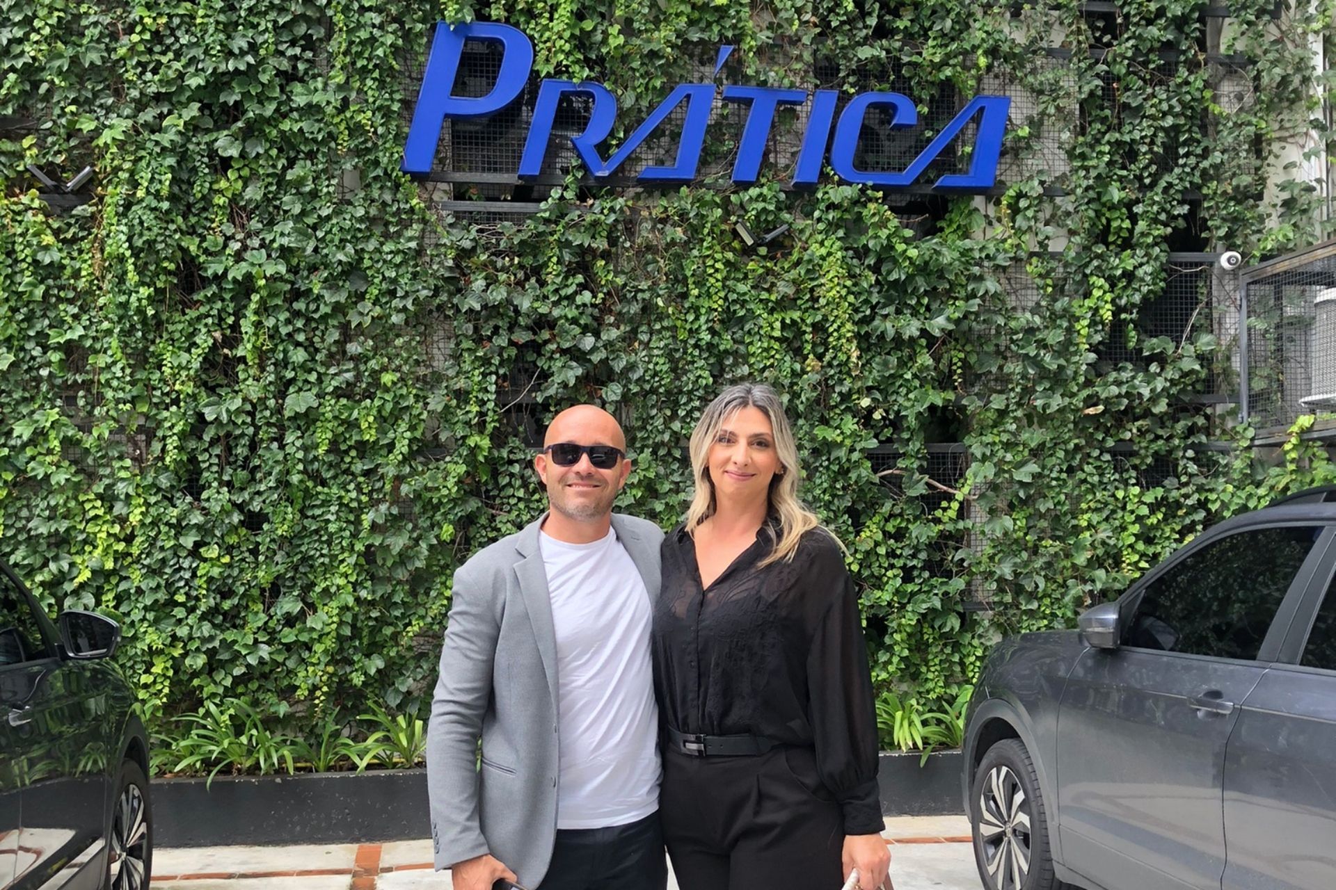 Duas pessoas posam em frente a uma parede coberta de hera e uma placa com o nome 