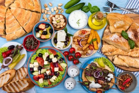 Uma seleção de aperitivos mediterrâneos, pão pita, azeitonas, pastas, saladas e petiscos fritos sobre uma toalha de mesa azul.