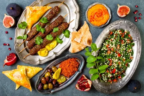 Pratos variados de comida halal, incluindo kebabs, saladas, molhos, figos e pão, dispostos em pratos ornamentados sobre uma mesa escura.