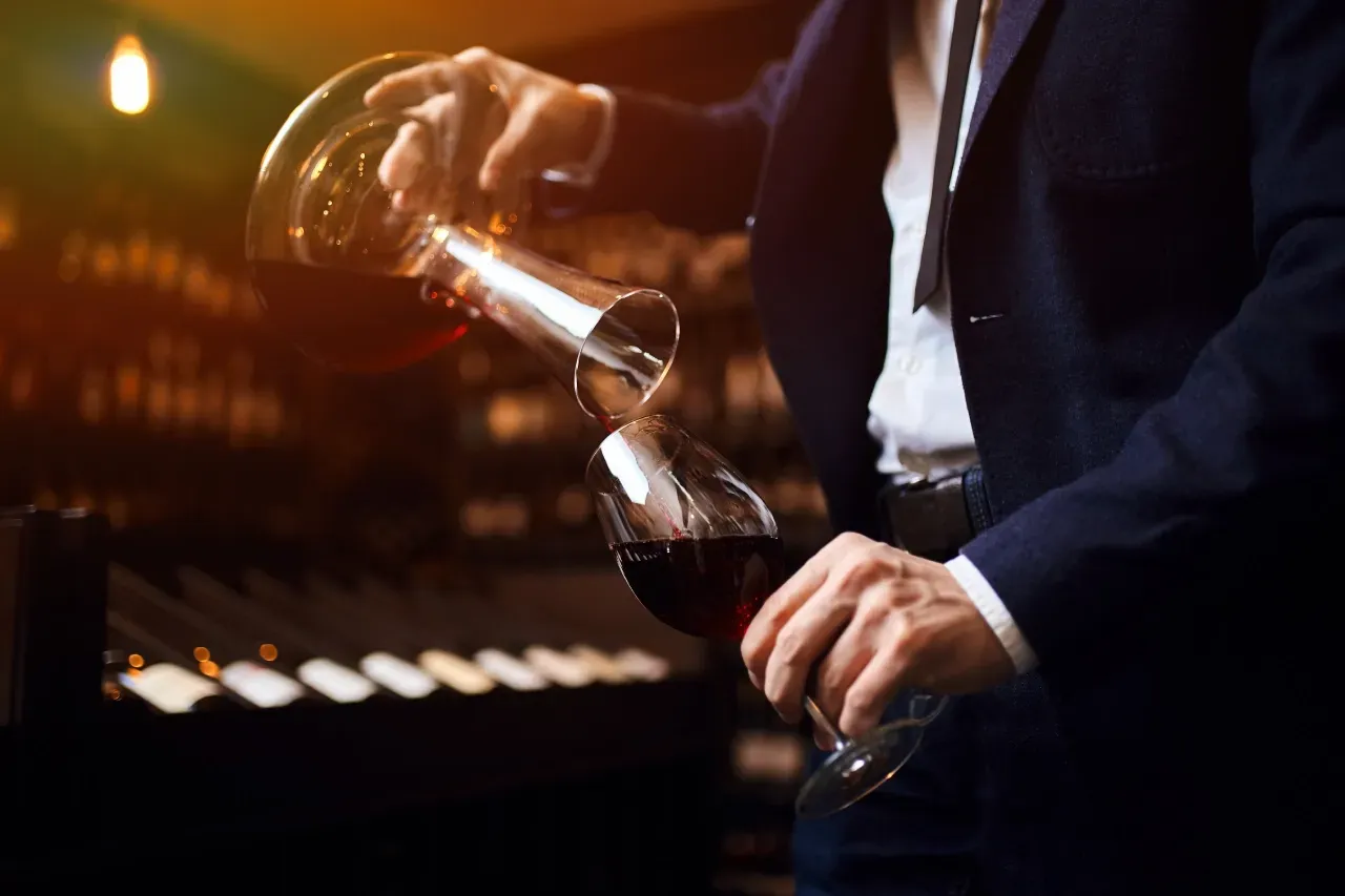 Pessoa de terno servindo vinho tinto de um decantador para uma taça em uma adega pouco iluminada.