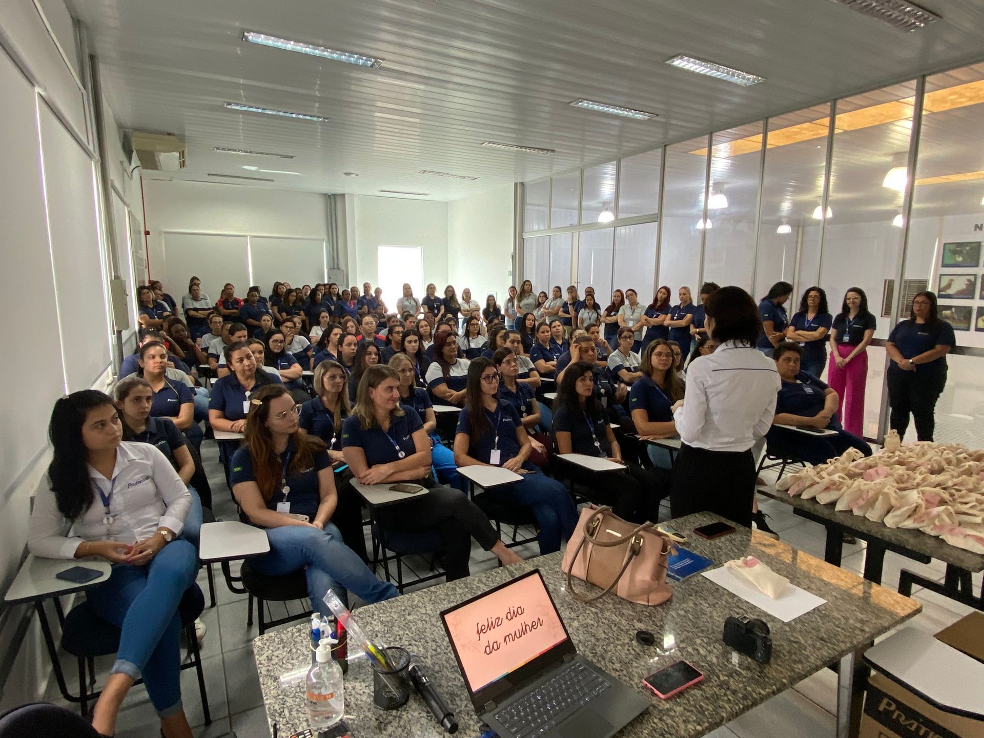 Um grande grupo de pessoas está sentado em carteiras em uma sala de aula.
