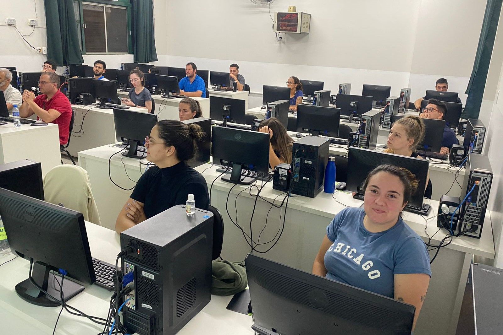Um grupo de pessoas está sentado em frente a monitores de computador em uma sala de aula.