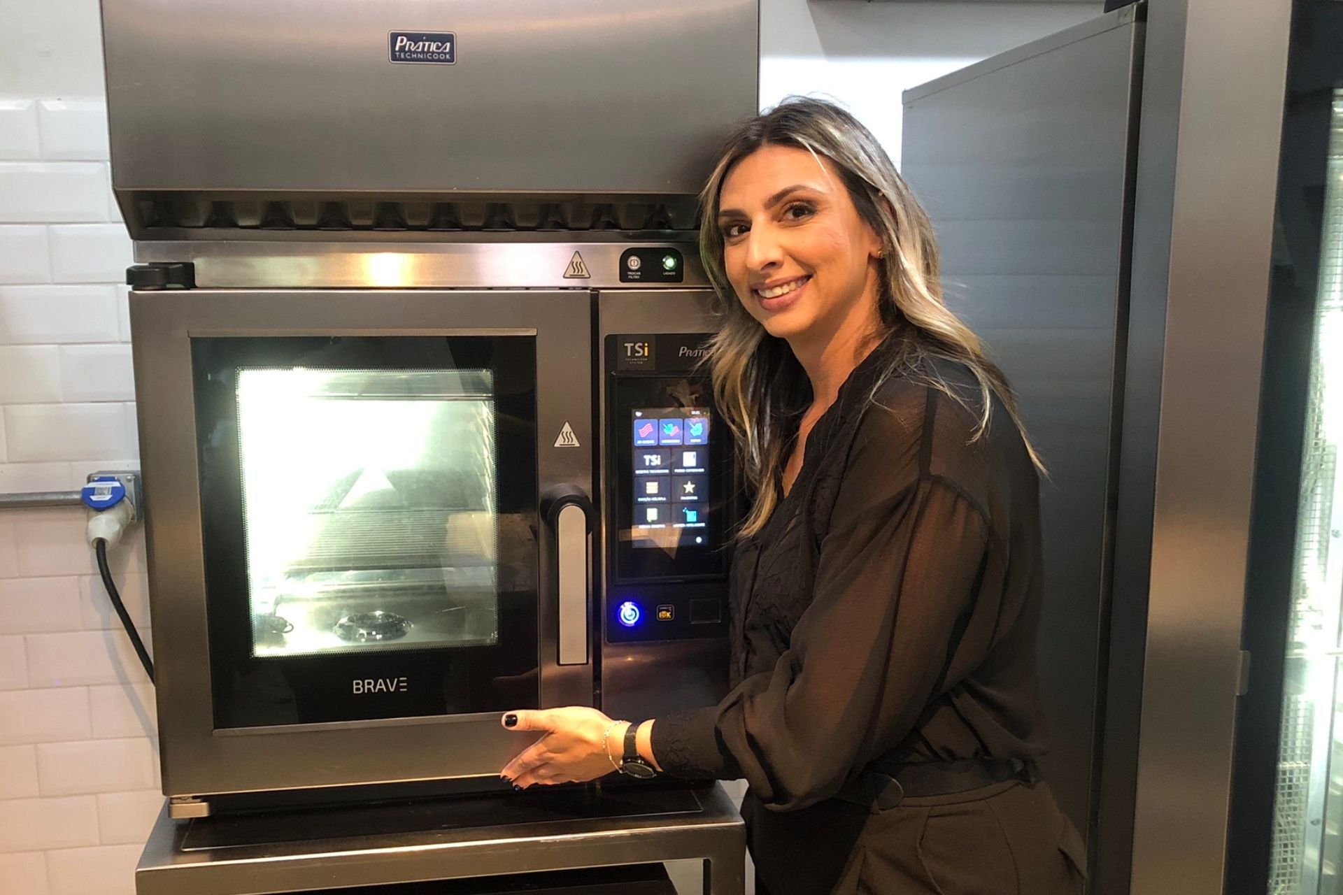 Mulher sorri ao abrir a porta de um forno industrial. Ambiente de cozinha industrial.