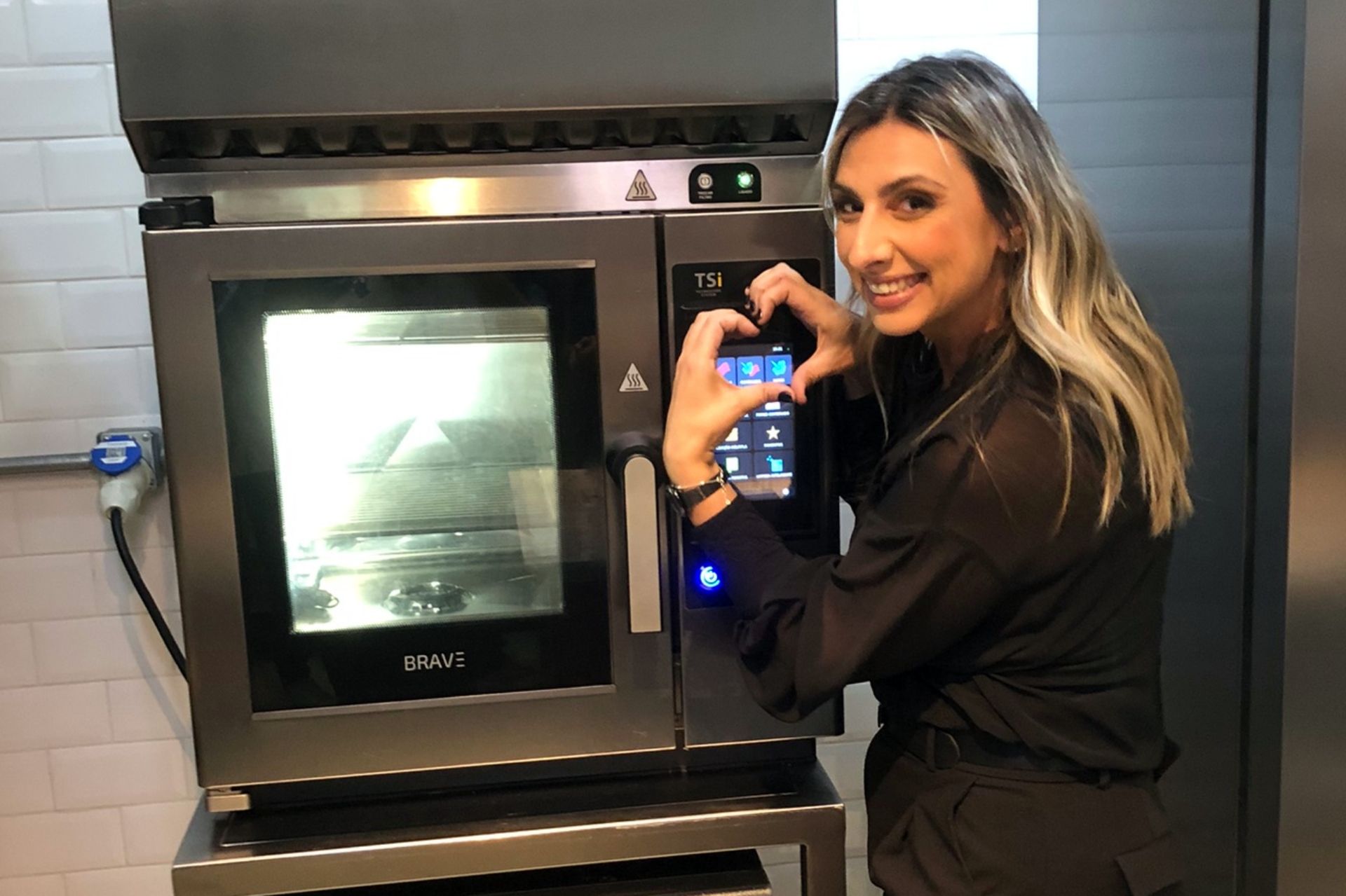 Mulher fazendo um coração com as mãos sobre um forno inteligente. Cozinha com acabamento em aço inoxidável e cores neutras.