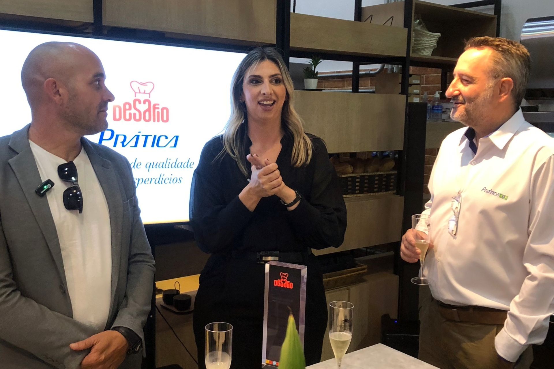 Três pessoas em um evento: mulher aplaudindo, homens segurando bebidas, logotipo na tela, ambiente interno.